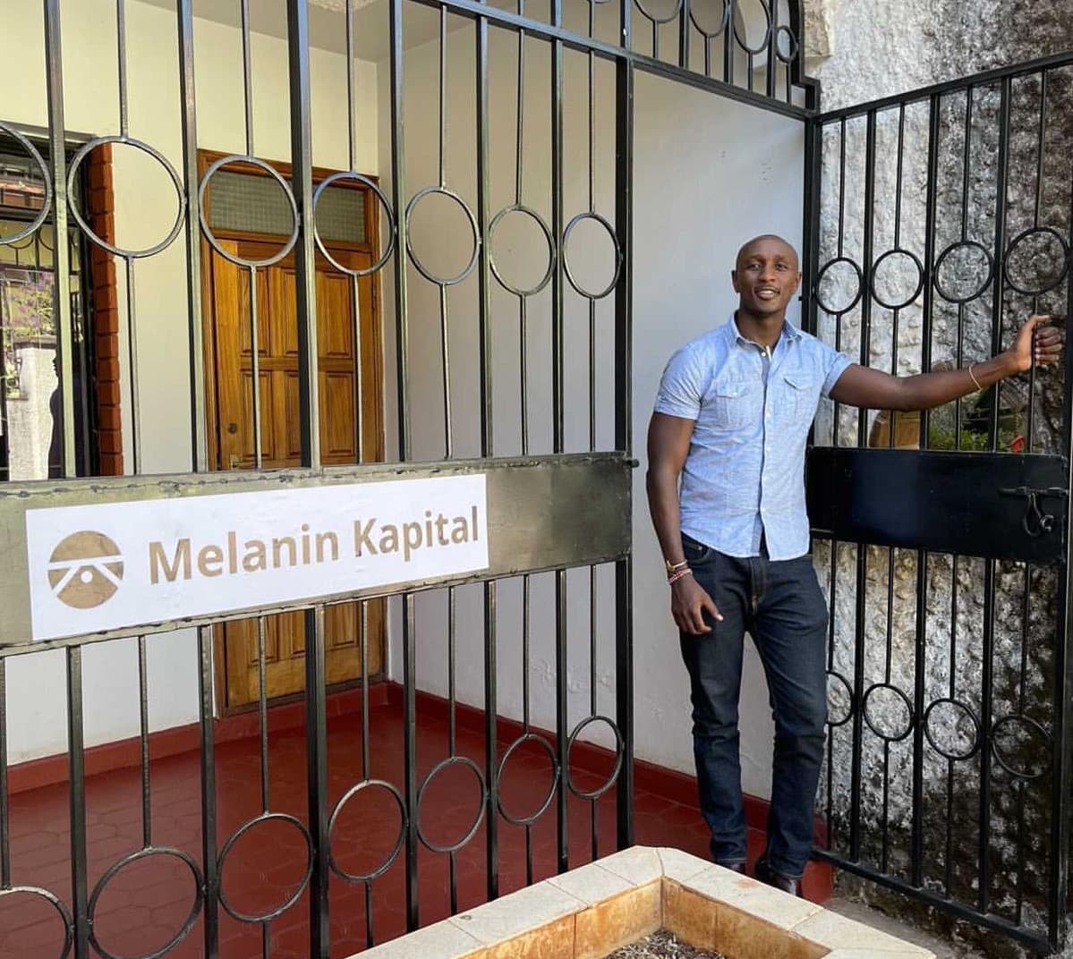 A member of the team at the gate of the Riverside Drive office, beside the Melanin Kapital sign